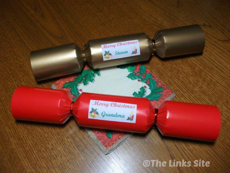 A red and a gold Christmas cracker on a wooden table with a Christmas serviette. The crackers have personalized labels for 'Steven' and 'Grandma'. 