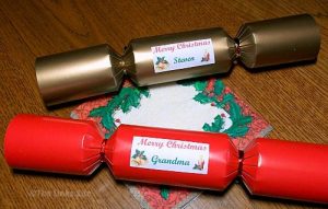 A red and a gold Christmas cracker on a wooden table with a Christmas serviette. The crackers have personalized labels for 'Steven' and 'Grandma'.