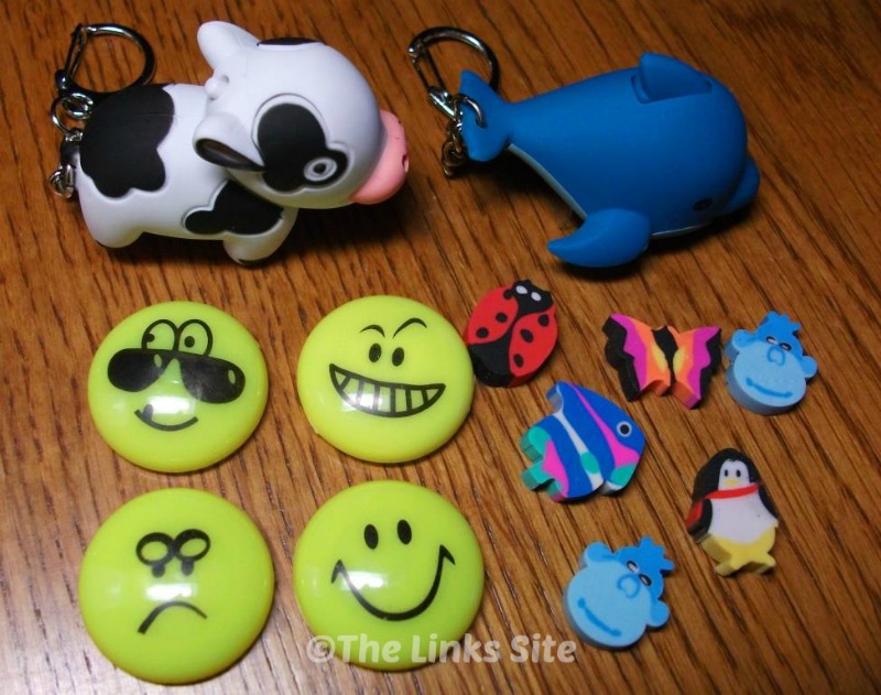 Various small items arranged on a wooden table. The items include; key-rings in the shape of a cow and a dolphin, emoji magnets, and small animal shaped erasers.