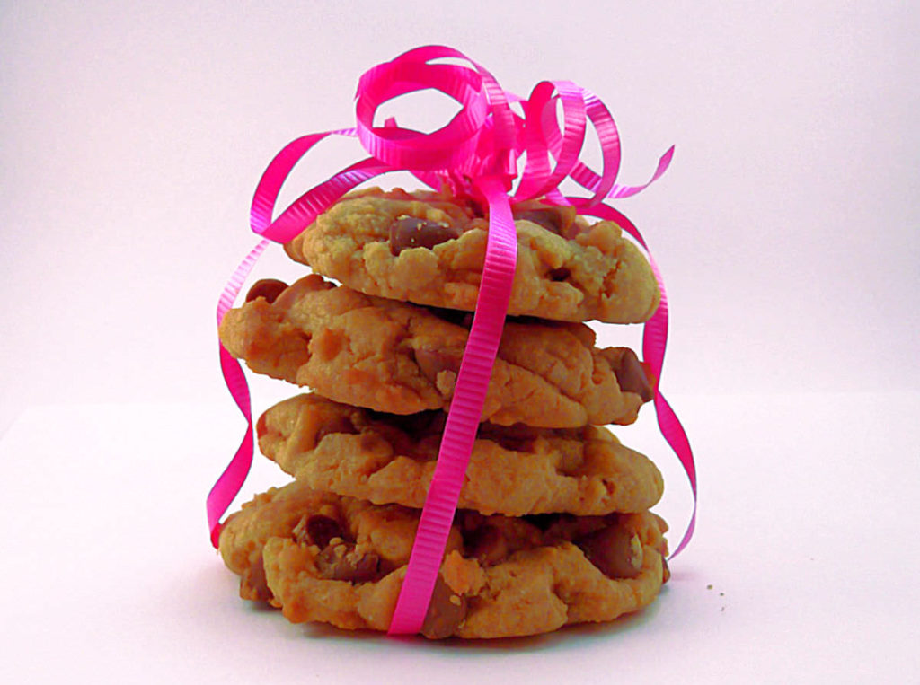 A stack of four chocolate chip cookies with a white background. The cookies are loosely wrapped with pink decorative curling ribbon and there is a bunch of curled ribbon on top of the cookies.