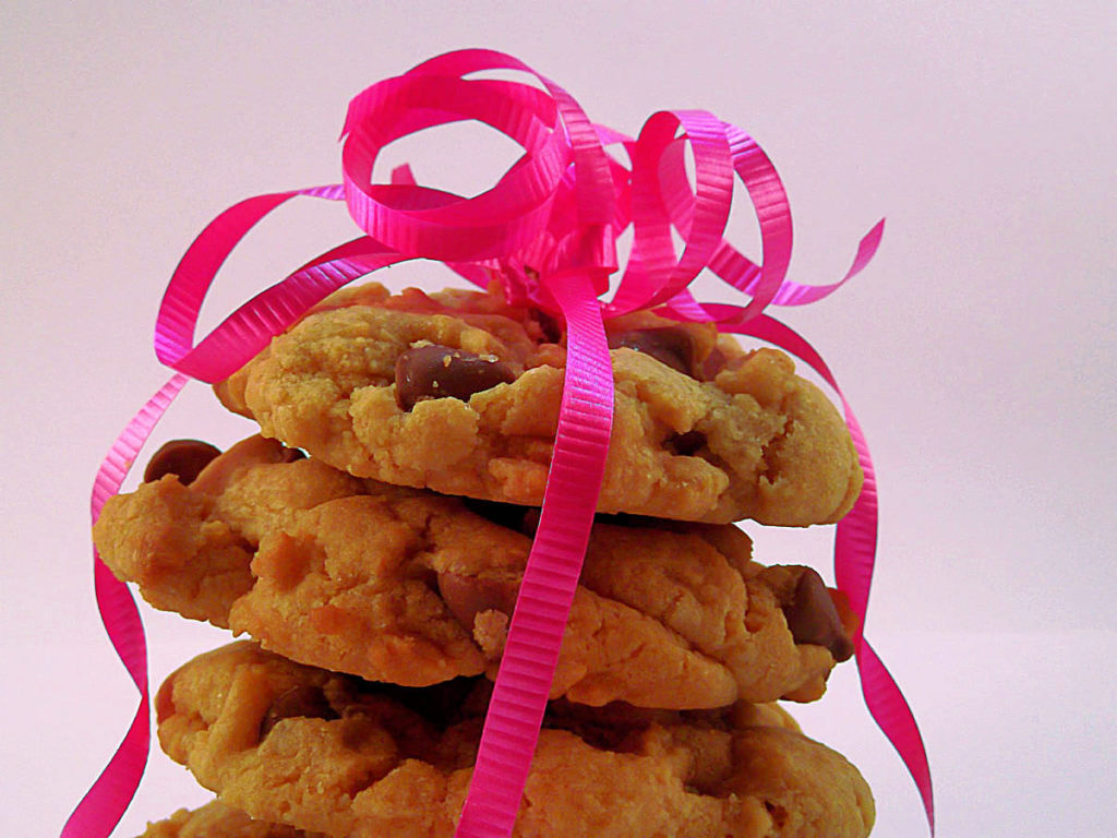 Close up picture of a stack of chocolate chip cookies with a white background. The cookies are loosely wrapped with pink decorative curling ribbon and there is a bunch of curled ribbon on top of the cookies.