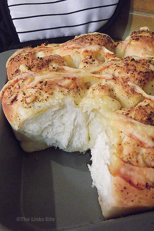 Baking dish containing cooked cheesy garlic and herb pull apart bread. A couple of piece of bread have been removed from the dish exposing the fluffy white bread. Black and white oven mitts can be seen in the background.