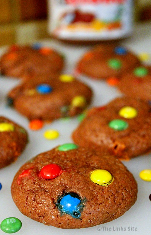 One cookie topped with M&M's is in focus in the foreground. More cookies and a jar of Nutella can be seen out of focus in the background.