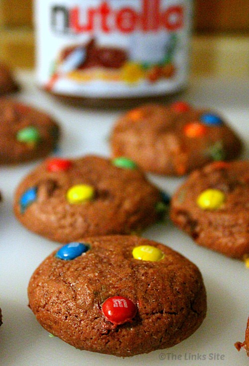 Plump, M&M topped Nutella cookies on a white kitchen board. A Nutella jar can be seen in the background.