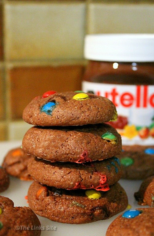Stack of 4 cookies on a white board with more cookies placed around the stack and a Nutella jar in the background.