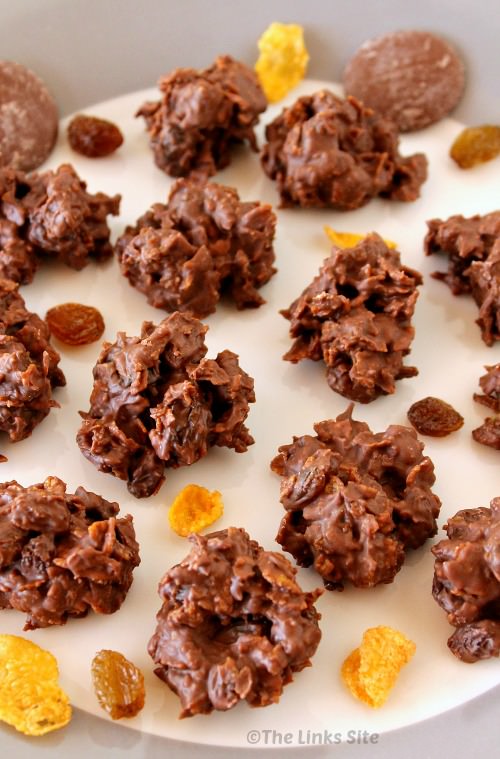 Several crunchy choc sultana bites are arranged on a grey and white plate. Some corn flake pieces, sultanas, and chocolate buttons are scattered around them