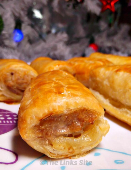 Close up of a sausage roll on a festive plate with other sausage rolls in the background. A Christmas tree is also in the background