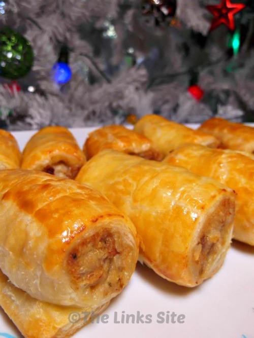 Two rows of small sausage rolls on a plate with a Christmas tree in the background.