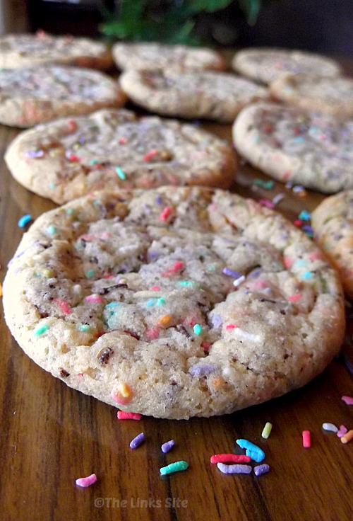 Several cookies are laid out on a wooden table with multi-coloured sprinkles scattered around them.