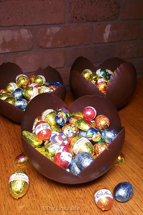3 Bowls made from chocolate and filled with small Easter eggs sitting on a wooden table.