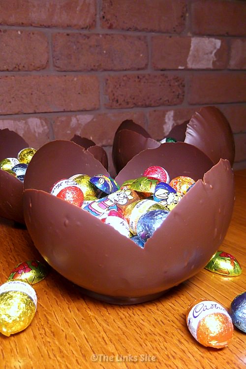 A bowl made from chocolate and filled with Easter chocolates is sitting on a wooden table. More Easter chocolates are scattered around on the table and two more chocolate bowls can be seen in the background.