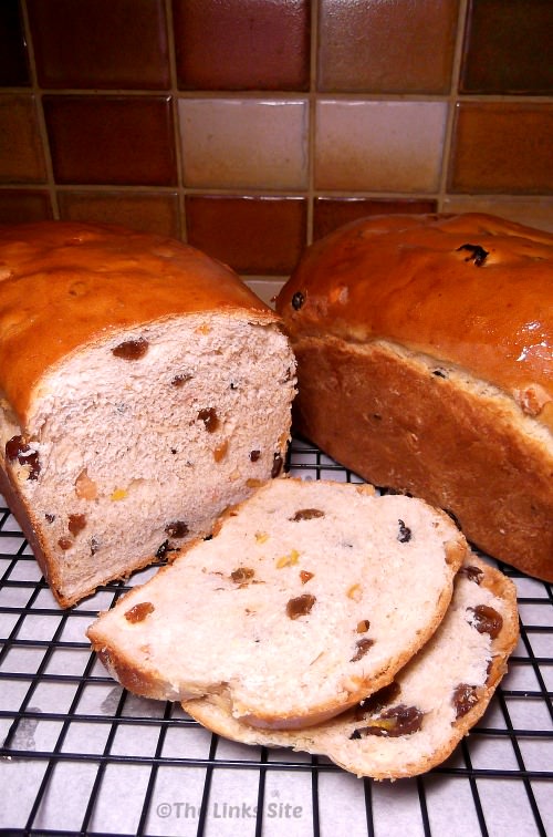 Two fruit loaves on a cooling rack, one of the loaves has been partially sliced. A tiled splashback in with brown colours can be seen in the background