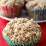 One apple crumble muffin in the foreground with a plate containing more muffins in the background.