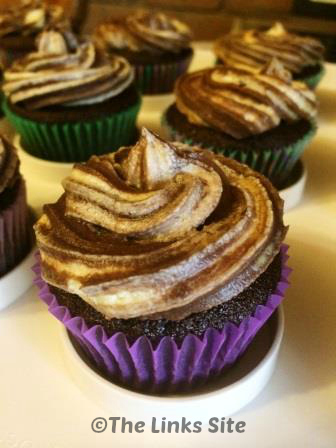 A choc orange cupcake in a purple paper case is pictured on white cupcake tray. It is topped with a swirl of marbled frosting and more cupcakes can be seen in the background.