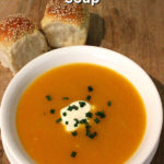 Overhead view of a white bowl filled with soup. The soup is topped with a small amount of sour cream along with some chopped chives. The bowl is placed on a wooden board along with two small bread rolls. Text overlay says: Quick & Easy Potato & Pumpkin Soup.