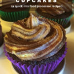 A choc orange cupcake in a purple paper case is pictured on white cupcake tray. It is topped with a swirl of marbled frosting and more cupcakes can be seen in the background. Text overlay says: Frosted Choc Orange Cupcakes (a quick mix food processor recipe!)