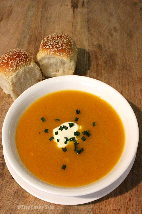 Overhead view of a white bowl filled with pumpkin soup. The soup is topped with a small amount of sour cream along with some chopped chives. The bowl is placed on a wooden board along with two small bread rolls.