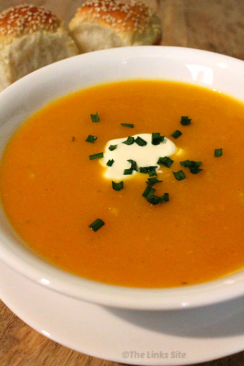 A white bowl filled with soup with a small amount of sour cream in the middle of the soup along with some chopped chives. Two sesame seed topped dinner rolls can be seen in the background.