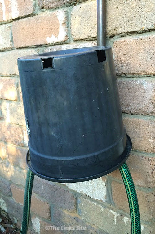 Garden tap that is attached to a brick wall. The garden tap is covered with an upturned black plastic plant pot. Two green garden hoses can be seen coming out from under the pot.