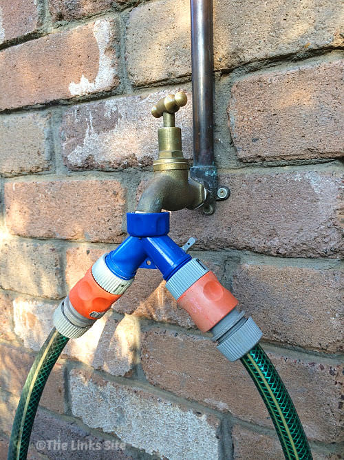 Garden tap attached to a brick wall. The tap is fitted with a plastic hose connector that acts as a two way adaptor so that two garden hoses can be connected to the one tap.