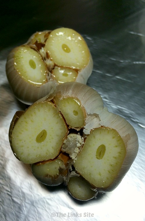 Two uncooked heads of garlic sitting on a piece of aluminium foil. The top of the head has been trimmed off and drizzled with oil.