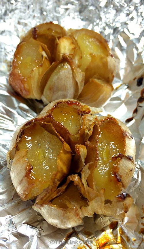 Two whole heads of roasted garlic with exposed cloves are sitting on a piece of crumpled aluminium foil.