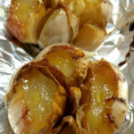 Two whole heads of roasted garlic with exposed cloves are sitting on a piece of crumpled aluminium foil. Text overlay says: How to Roast Garlic in the Oven.
