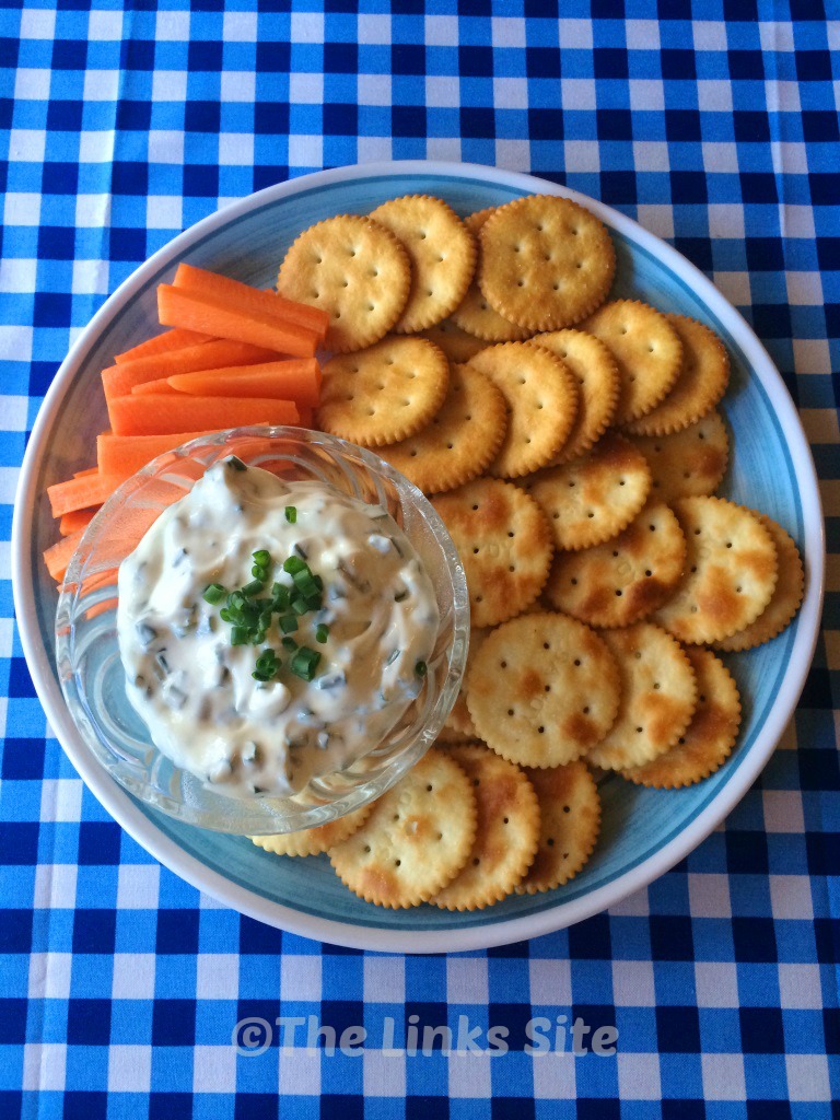 Overhead view of a blue and white plate which contains crackers, carrot sticks, and a glass blow of dip. The plate is placed on a blue and white check tablecloth. 