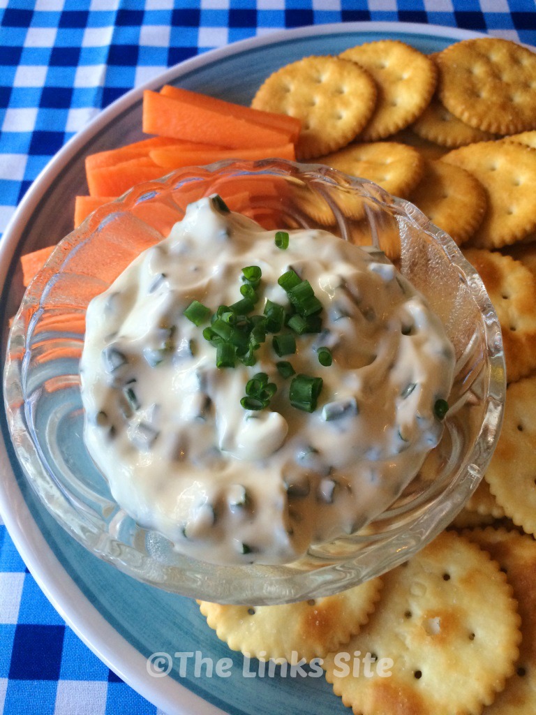 A glass bowl containing dip is placed on a blue and white plate. Carrot sticks and crackers are also placed on the plate.
