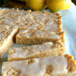 Small pieces of slice are pictured on a piece of baking paper with some whole lemons in the background. Text overlay says: Tangy Lemon Coconut Slice (low calorie & no bake!).