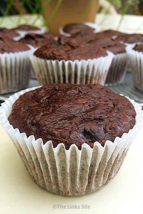One chocolate muffin is place in the foreground and several other muffins on a silver plate can be seen in the background.