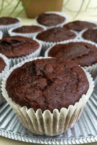 A chocolate muffin in a white paper case on a silver plate with several more muffins behind it.