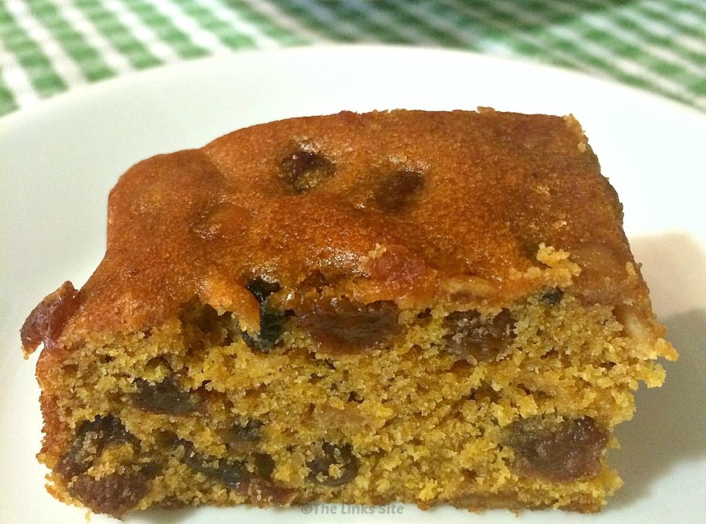 A single piece of pumpkin fruit cake on a white plate. The white plate is sitting on a green and white check tea towel.