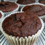 A chocolate muffin in a white paper case on a silver plate with several more muffins behind it. Text overlay says: Double Chocolate Sour Cream Muffins.