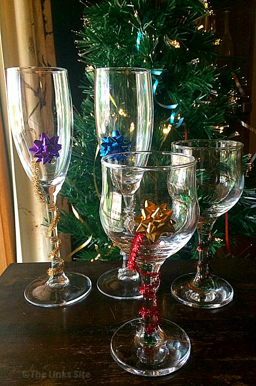 Four decorated wine glasses on a wooden table with a Christmas tree in the background.