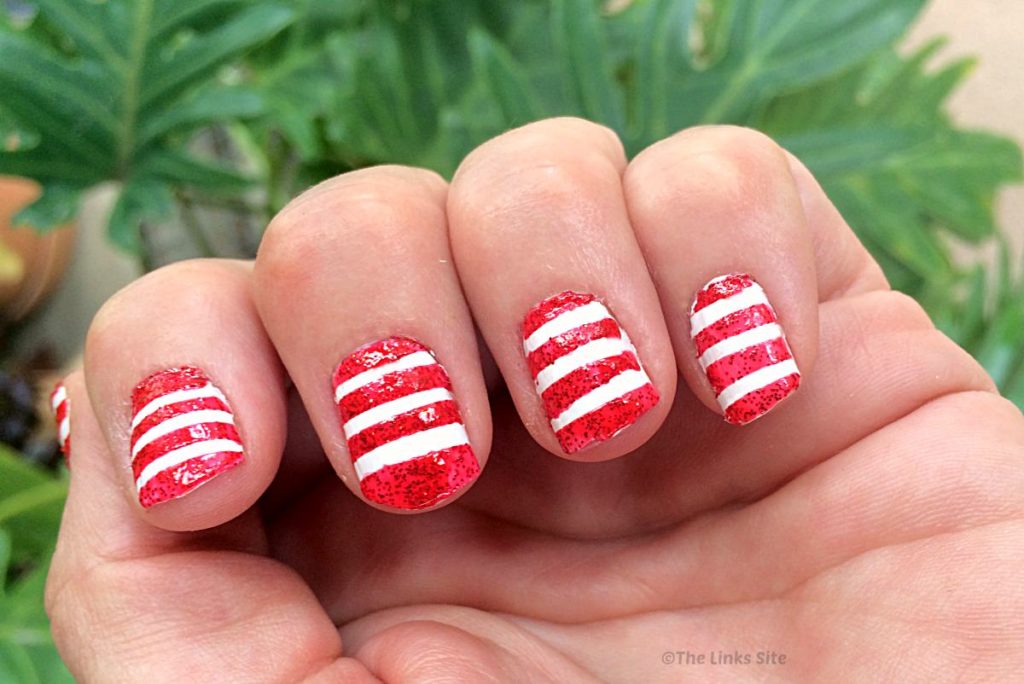 Wide image showing four fingers on a hand that have been painted with nail polish in a red an white stripy pattern. Plants can be seen in the background.