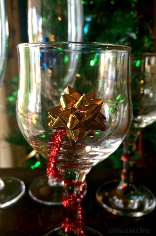 Close up of a wine glass that is decorated with a red pipe cleaner and a small gold gift bow.
