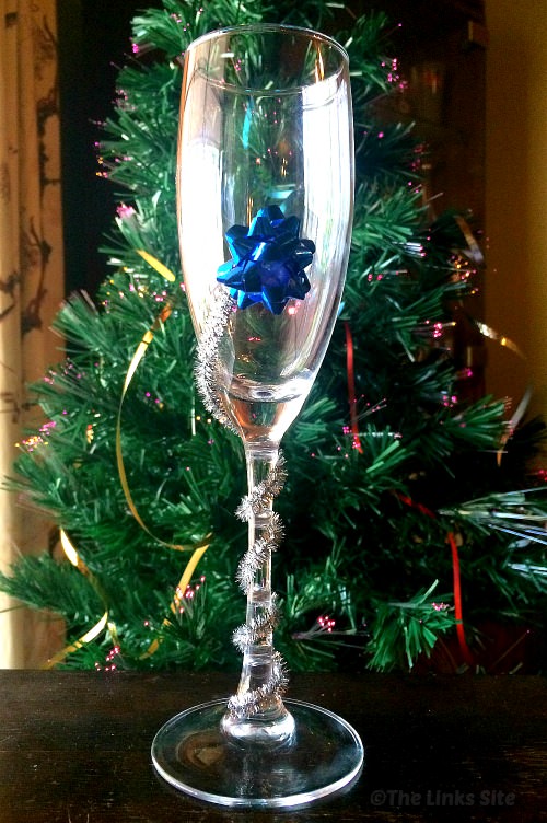 A single long stem champagne glass decorated with a silver pipe cleaner and a blue gift bow. A Christmas tree can be seen in the background.