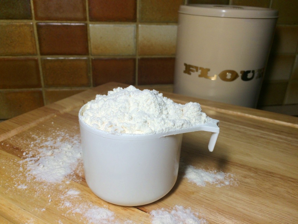 White measuring cup filled with flour is placed on a wooden cutting board. There is flour scattered around the measuring cup and a storage canister labelled flour in the background.
