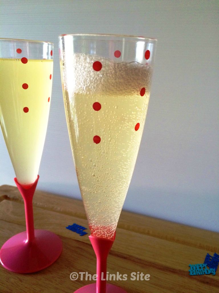 A plastic glasses of bubbly mock champagne on a wooden bench. The base and the stem of the glass are pink while the top is clear with pink dots. Another glass can be seen in the background.