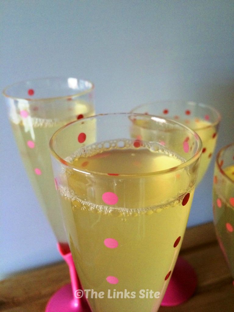 Close up of a glass of mock champagne. The glass is clear with pink spots and other glasses can be seen in the background.