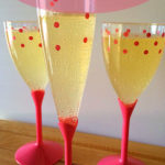 Three plastic glasses of mock champagne on a wooden bench. The base and the stem of the glasses are pink while the tops are clear with pink dots. Text overlay says: Easy 2 ingredient Kids Champagne.