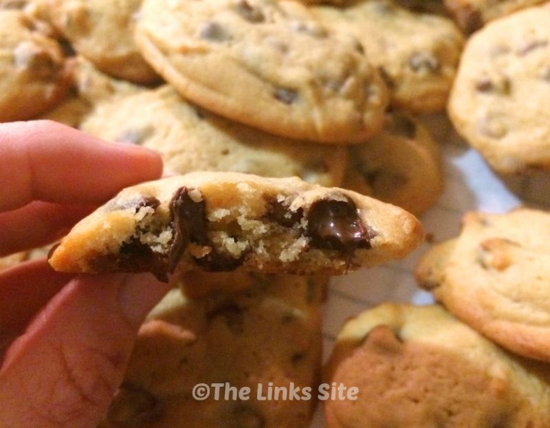 One cookie that is still warm has been broken open and is being held up to the camera to show soft gooey chocolate and caramel chips. A plate with more cookies can be seen in the background.