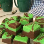 Squares of green and brown fudge on a white plate. A spoon, two dark green coffee mugs, and a blue and white check tea towel can be seen in the background.