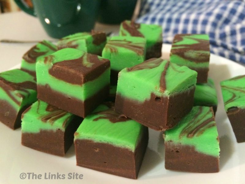 Squares of green and brown fudge on a white plate. Dark green coffee mugs and a blue and white check tea towel can be seen in the background.