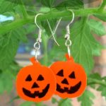 Orange Jack O Lantern shaped earrings are pictured hanging from plant material. Text overlay says: Cute Recycled Plastic Halloween Earrings