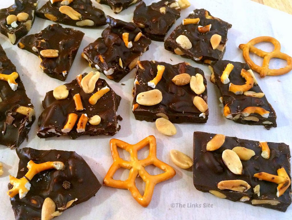 Roughly broken up pieces of chocolate bark arranged on baking paper. Mini pretzels and peanuts are also scattered around.