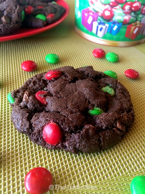 A cookie on a baking mat with red and green M&M's scattered around it. A red plate with more cookies as well as a bucket of M&M's can be seen in the background.
