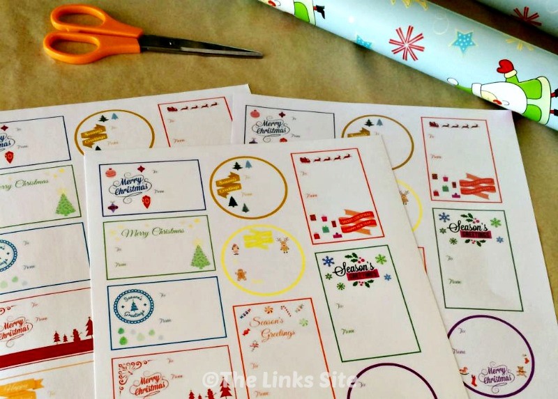Three sheets of gift tags on a craft table. Christmas wrapping paper and a pair of scissors can also be seen.