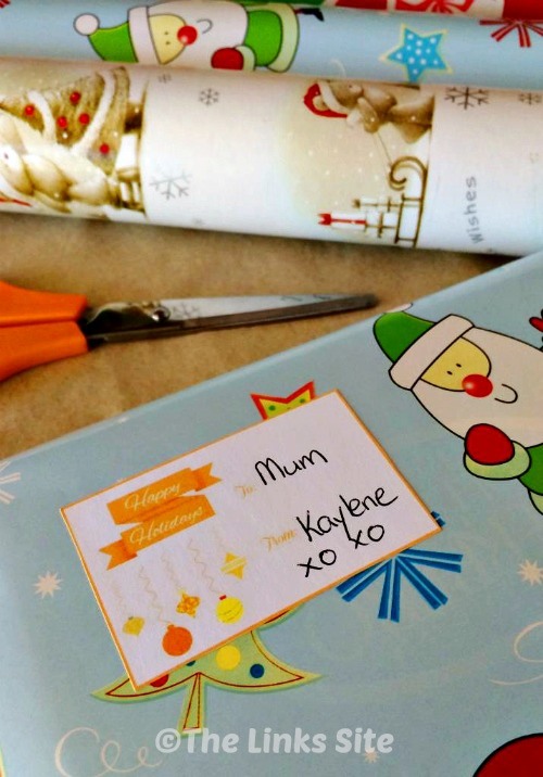 Close up of a gift tag on a wrapped present. Scissors and rolls of wrapping paper can be seen in the background.